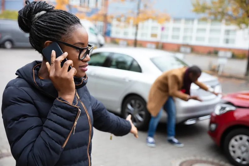 A woman on her phone at the roadside after a collision, with another person leaning over the damaged car behind her
