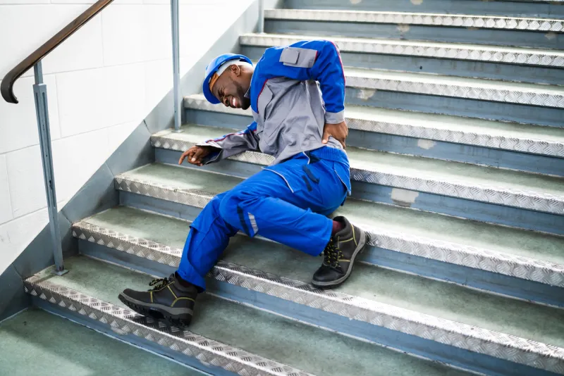 A worker in blue coveralls and a hard hat fallen on a concrete stairwell, holding his back