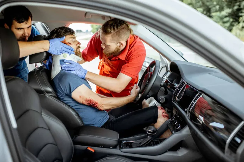 Rescuers fit a neck brace on an injured driver with a bleeding arm, still seated behind the wheel