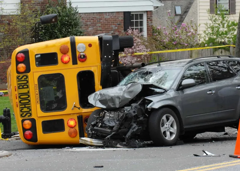Grey SUV with a crushed front end beside a yellow school bus lying on its side after a crash