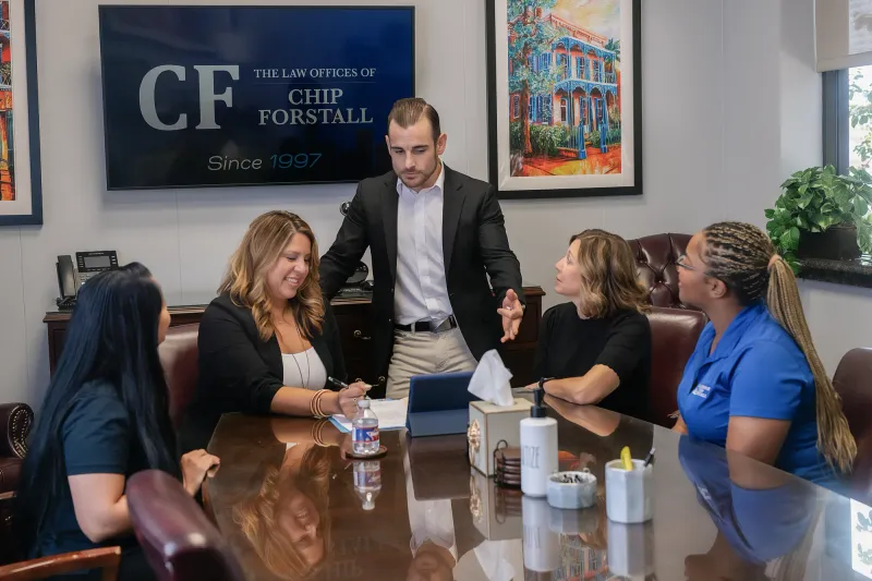 Jonathan Forstall standing at the head of the conference table with four colleagues seated around it, under the Law Offices of Chip Forstall sign